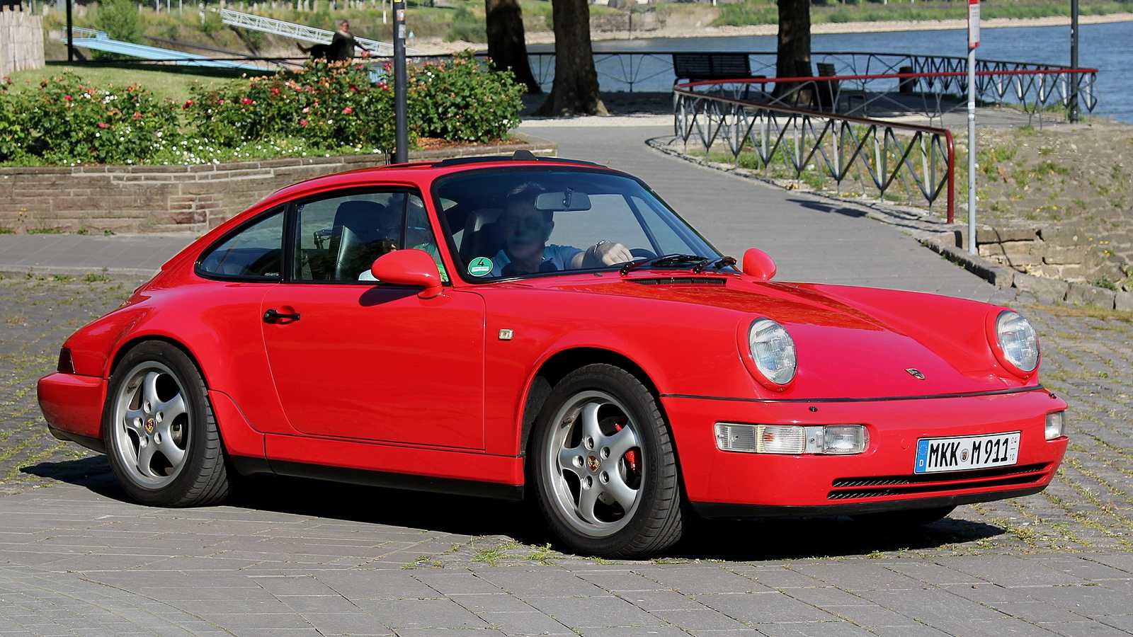 A red Porsche 911 Carrera of the 964 generation parked beside a river promenade