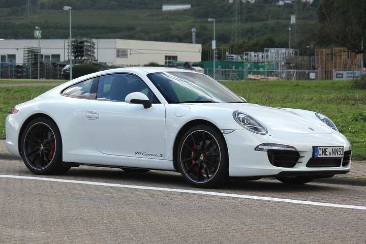 A white Porsche 911 Carrera S of the 991 generation driving past a field